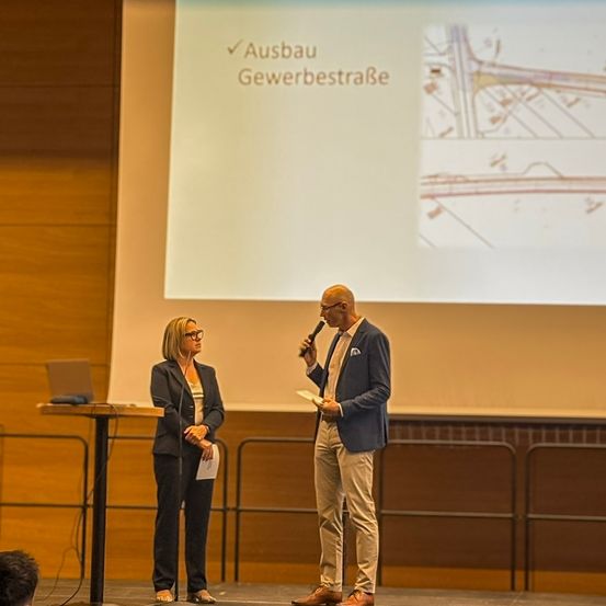 Ein Mann und eine Frau stehen mit Mikrofon und Papieren auf der Bühne. Eine Projektionsleinwand hinter ihnen zeigt ein Bild mit deutschem Text.