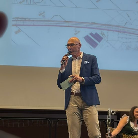 Ein Mann hält eine Rede auf der Bühne und hat ein Mikrofon in der Hand. Eine Frau spielt im Hintergrund ein Musikinstrument.