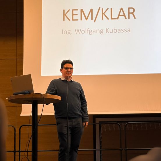 Ein Mann steht an einem Podium mit einem Mikrofon in einem Konferenzraum mit einer Projektionsleinwand dahinter, auf der KER/KLAR Ing. Wolfgang Kubasa steht.
