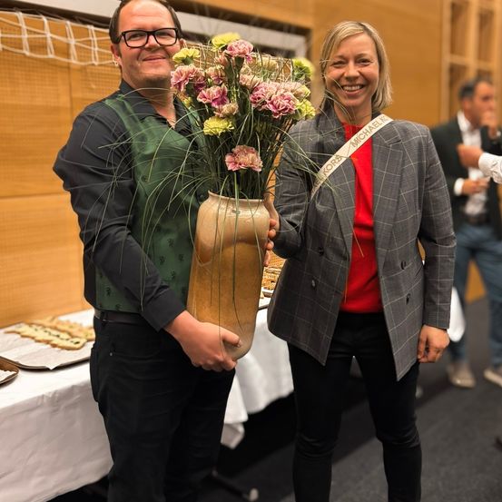 Ein Mann und eine Frau stehen lächelnd nebeneinander und halten einen großen Blumenstrauß. Die Frau trägt eine Schärpe mit der Aufschrift Michael Kobra. Hinter ihnen ist ein Tisch mit Essen und anderen Personen zu sehen.