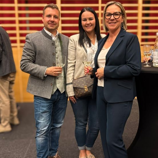 Drei Personen stehen lächelnd eng beieinander bei einer Veranstaltung. Ein Mann in einer grauen Jacke und Jeans hält ein Weinglas. Eine Frau in einem weißen Hemd und Jeans hält ebenfalls ein Glas. Eine weitere Frau in einem marineblauen Anzug hält ein Glas und trägt eine Brille. Hinter ihnen steht eine Holzwand und ein Tisch mit Weinflaschen.