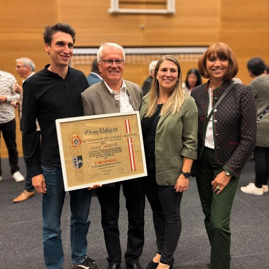 Vier Personen posieren für ein Bild, während sie ein Zertifikat in einem formellen Rahmen halten. Der Mann links trägt eine Brille, und die Frau rechts lächelt. Die Frau in der Mitte hält das Zertifikat mit beiden Händen. Der Mann rechts lächelt und trägt einen Blazer.