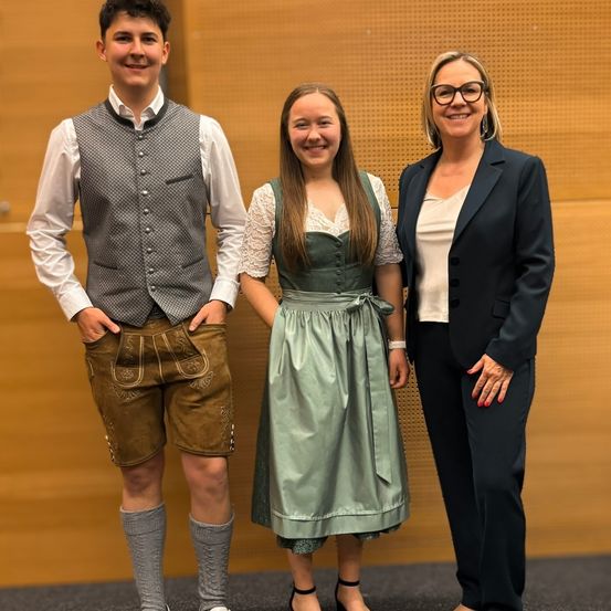 Drei Menschen stehen eng beieinander und lächeln. Der Mann trägt eine Weste mit Knöpfen und eine kurze Hose. Die Frau in der Mitte trägt ein Kleid mit einem Gürtel. Die andere Frau trägt eine Brille und einen Anzug.