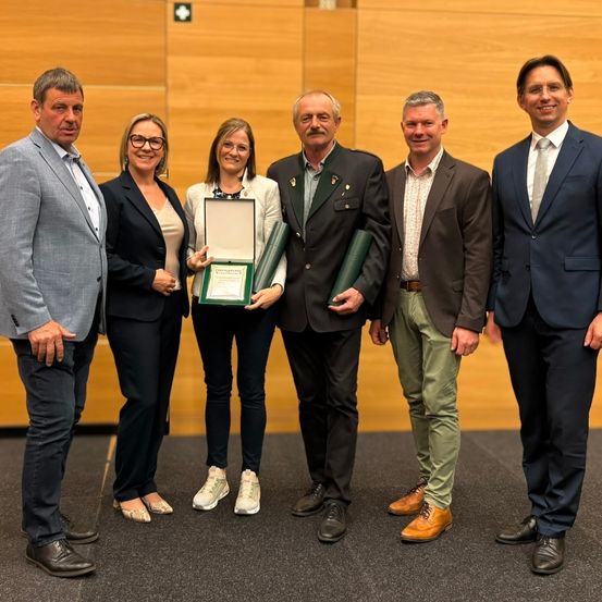 Fünf Personen in formeller Kleidung stehen lächelnd zusammen in einem Innenraum. Eine Frau in der Mitte hält einen gerahmten Preis und zwei Männer neben ihr halten grüne Ordner. Dahinter befindet sich eine Holzwand.
