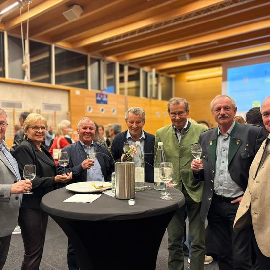 Eine Gruppe älterer Männer und Frauen steht um einen Tisch mit Weinläsern und Weinflaschen. Sie lächeln und posieren für ein Foto. Der Tisch hat einen Teller mit Essen, eine Flasche und einen silbernen Eimer. Dahinter ist ein verschwommener Blick auf einen Raum mit Holzwänden und einer Projektionsleinwand.