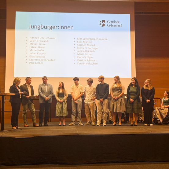 Eine Gruppe von Menschen steht auf einer Bühne mit einer Präsentation im Hintergrund. Die Präsentation zeigt Namen von Personen, möglicherweise Teilnehmer eines Events.