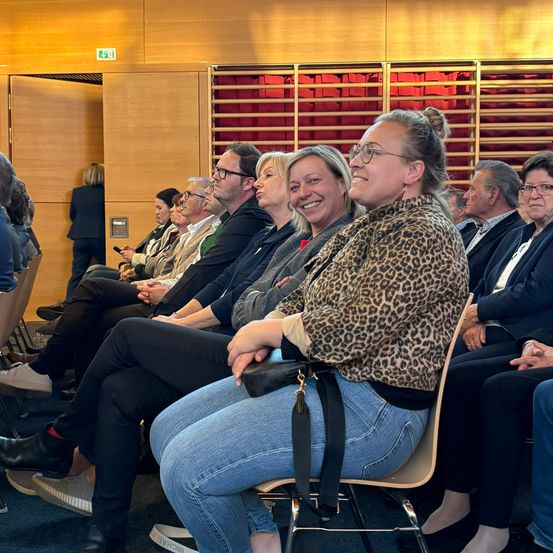 Eine Gruppe von Menschen sitzt in einem Auditorium. Eine Frau in einer Leoparden-Jacke lächelt und schaut in die Kamera. Andere sitzen vor ihr. Eine Person steht hinten.