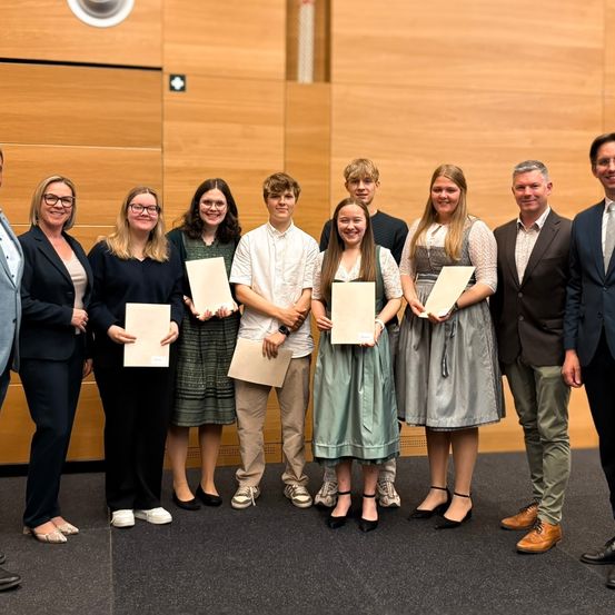 Eine Gruppe von Menschen, einige mit Urkunden in der Hand, steht in einem formellen Rahmen zusammen. Sie sind in professioneller Kleidung gekleidet und scheinen für ein Foto zu posieren.
