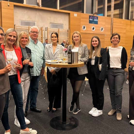 Eine Gruppe von Menschen versammelt sich um einen Tisch in einem Raum, einige halten Weingläser in den Händen. Eine Frau mit einer Handtasche steht neben einem Tisch mit Flaschen und Gläsern. Dahinter befindet sich eine Wand mit einem Schild.