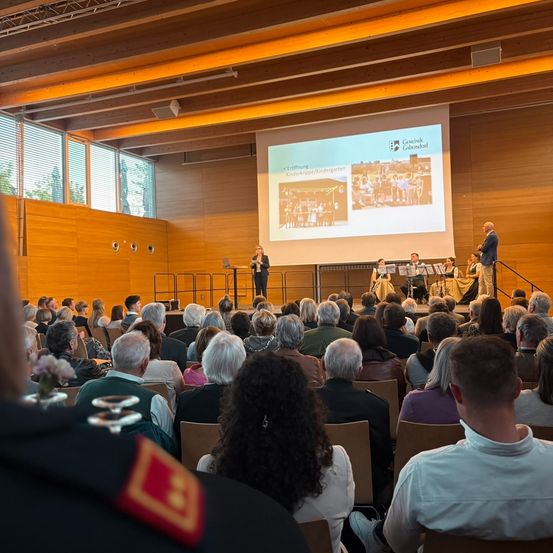 Eine Vorlesung findet in einem großen Hörsaal statt. Eine Frau hält eine Rede am Rednerpult. Hinter ihr zeigt eine Leinwand Bilder. Das Publikum sitzt in Reihen und hört aufmerksam zu.