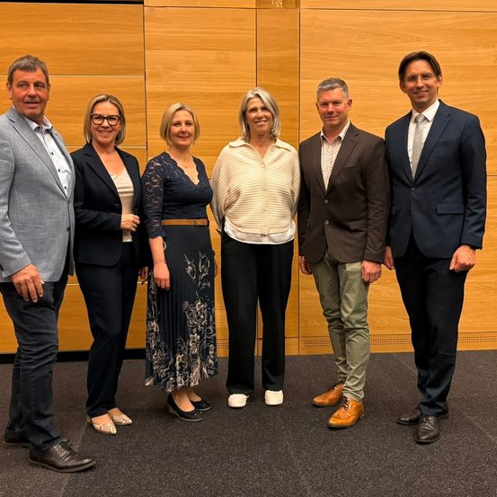 Sechs Menschen stehen nebeneinander und lächeln für ein Foto. Eine Frau in einem weißen Oberteil steht in der Mitte. Sie sind alle formell gekleidet, Männer in Anzügen und Frauen in Abendkleidern. Hinter ihnen befindet sich eine Holzwand.