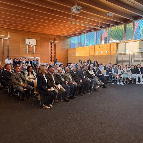 Eine Gruppe von Menschen sitzt in einem großen Innenraum, einige sind formell gekleidet, andere in Freizeitkleidung. Der Raum hat eine Holzdecke, Wände und Boden. An der Wand hängt ein Basketballkorb.