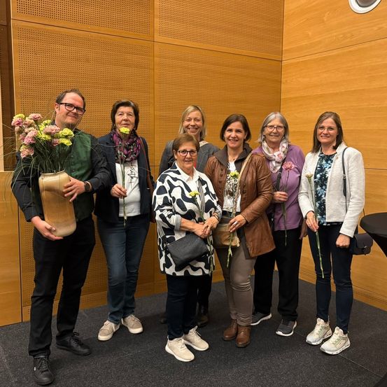 Sieben Personen lächeln in einem Raum mit einer Holzwand. Ein Mann hält eine Vase mit Blumen, während die Frauen Blumen in den Händen haben. Sie tragen alle Freizeitkleidung.