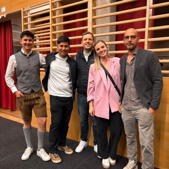 Fünf Menschen, vier Männer und eine Frau, stehen dicht beieinander vor einer Holzwand, lächeln und posieren für ein Foto.