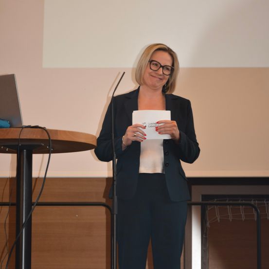 Eine Frau hält ein Papier in der Hand und steht vor einem Mikrofon an einem Podium vor einer weißen Leinwand.