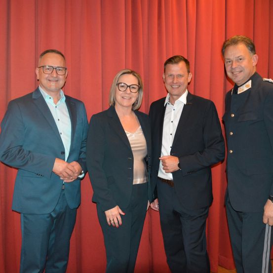 Vier Personen, zwei Männer und zwei Frauen, in formeller Kleidung, stehen vor einem roten Vorhang. Sie scheinen zu lächeln und posieren für ein Foto.