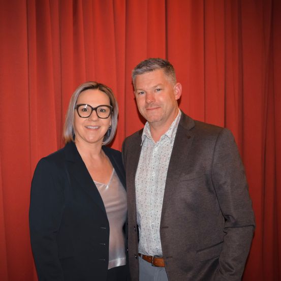 Ein Mann und eine Frau stehen nebeneinander vor einem roten Vorhang. Die Frau trägt eine Brille und einen schwarzen Blazer und lächelt. Der Mann trägt einen braunen Blazer mit einem weißen Hemd und lächelt auch.