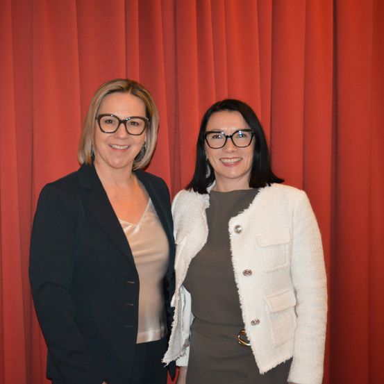 Zwei Frauen mit Brille stehen lächelnd vor einem roten Vorhang. Eine trägt einen schwarzen Blazer, die andere eine weiße Jacke.
