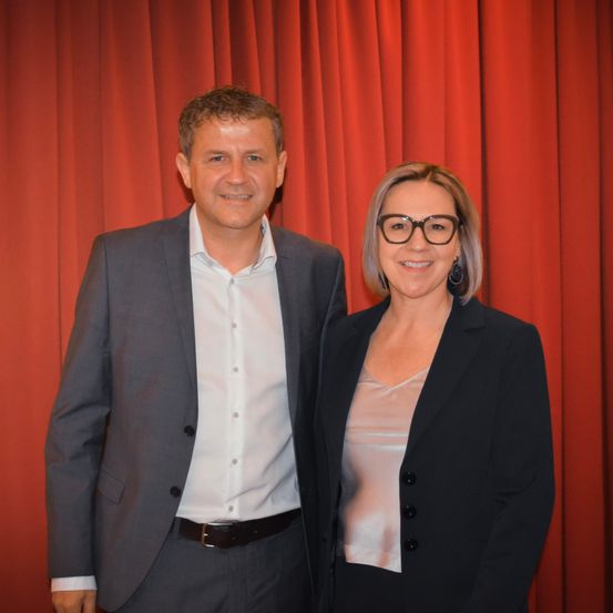 Ein Mann und eine Frau stehen lächelnd nebeneinander vor einem roten Vorhang. Beide tragen beruflich gekleidet.