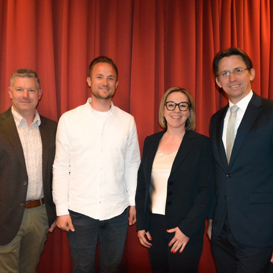 Vier Personen, zwei Männer und zwei Frauen, stehen gemeinsam vor roten Vorhängen. Sie sind in formeller Kleidung gekleidet und lächeln für ein Foto.