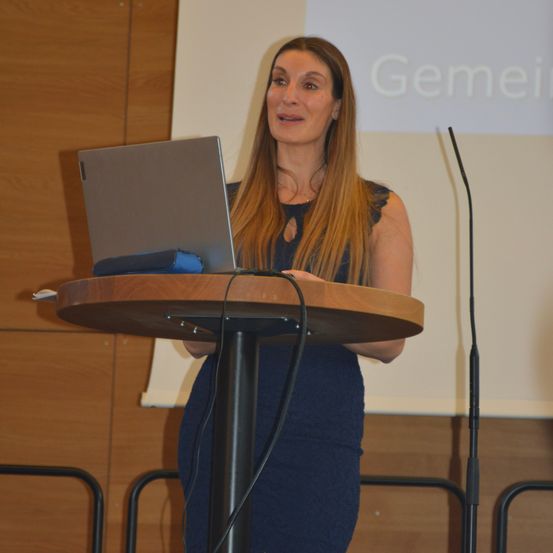 Eine Frau in einem blauen Kleid steht an einem Rednerpult mit einem Laptop vor einem Publikum. Ein Mikrofon und eine Projektionsleinwand mit der Aufschrift 'Gemein' sind hinter ihr.