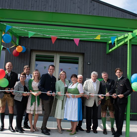 Eine Gruppe von Menschen, darunter Männer und Frauen, steht vor einem Gebäude und hält ein Band. Sie lächeln und scheinen ein Ereignis zu feiern. Das Gebäude hat ein grünes Dach mit Wimpelgirlande. Einige von ihnen tragen Brillen und einige tragen Hüte. Es gibt Ballons und Topfpflanzen auf dem Boden.