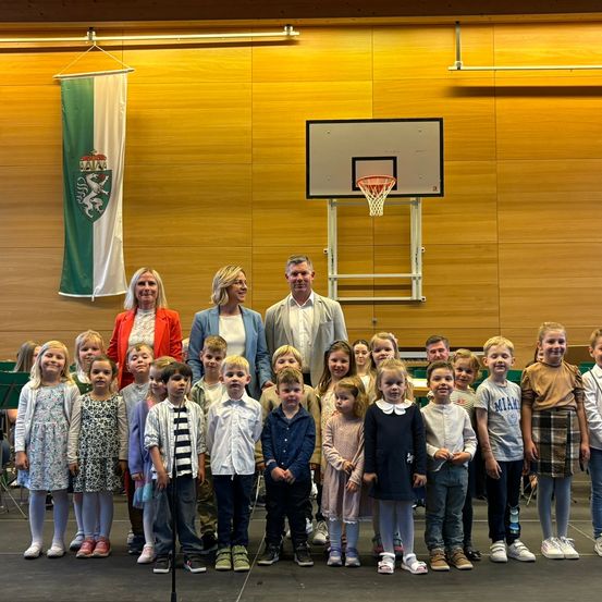 Eine Gruppe von Kindern und Erwachsenen steht auf einer Bühne mit einem Basketballkorb im Hintergrund. Zwei Frauen und ein Mann stehen vorne, und eine Fahne hängt an der Wand.