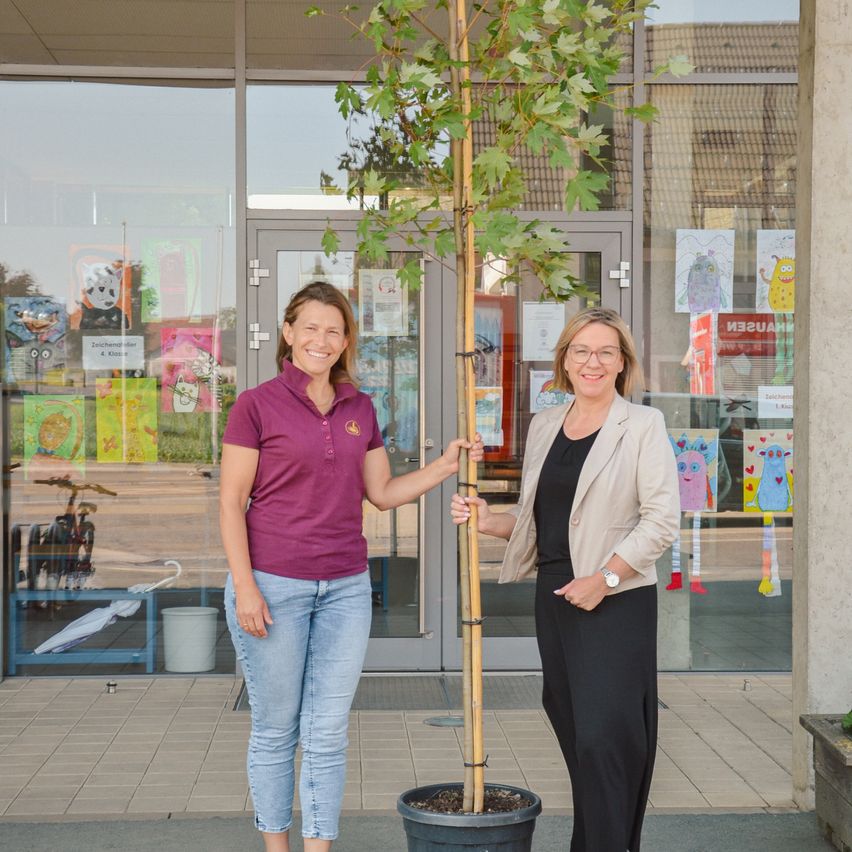Zwei Frauen stehen vor einem Gebäude mit einem Baum. Eine trägt ein lila Hemd, die andere eine beige Blazer. Sie lächeln beide für die Kamera.