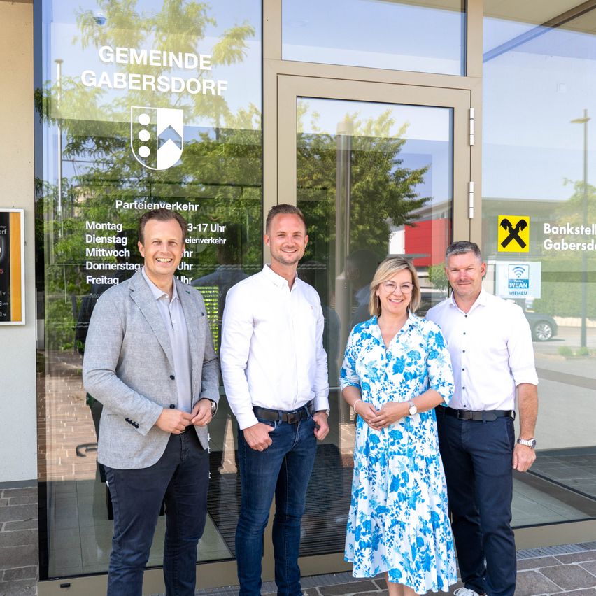 Drei Personen, zwei Männer und eine Frau, stehen vor dem Gemeindebüro Gabersdorf. Sie lächeln alle und scheinen für ein Foto zu posieren. Hinter ihnen ist der Eingang des Büros zu sehen, der mit einer Glastür und einem Logo gekennzeichnet ist.