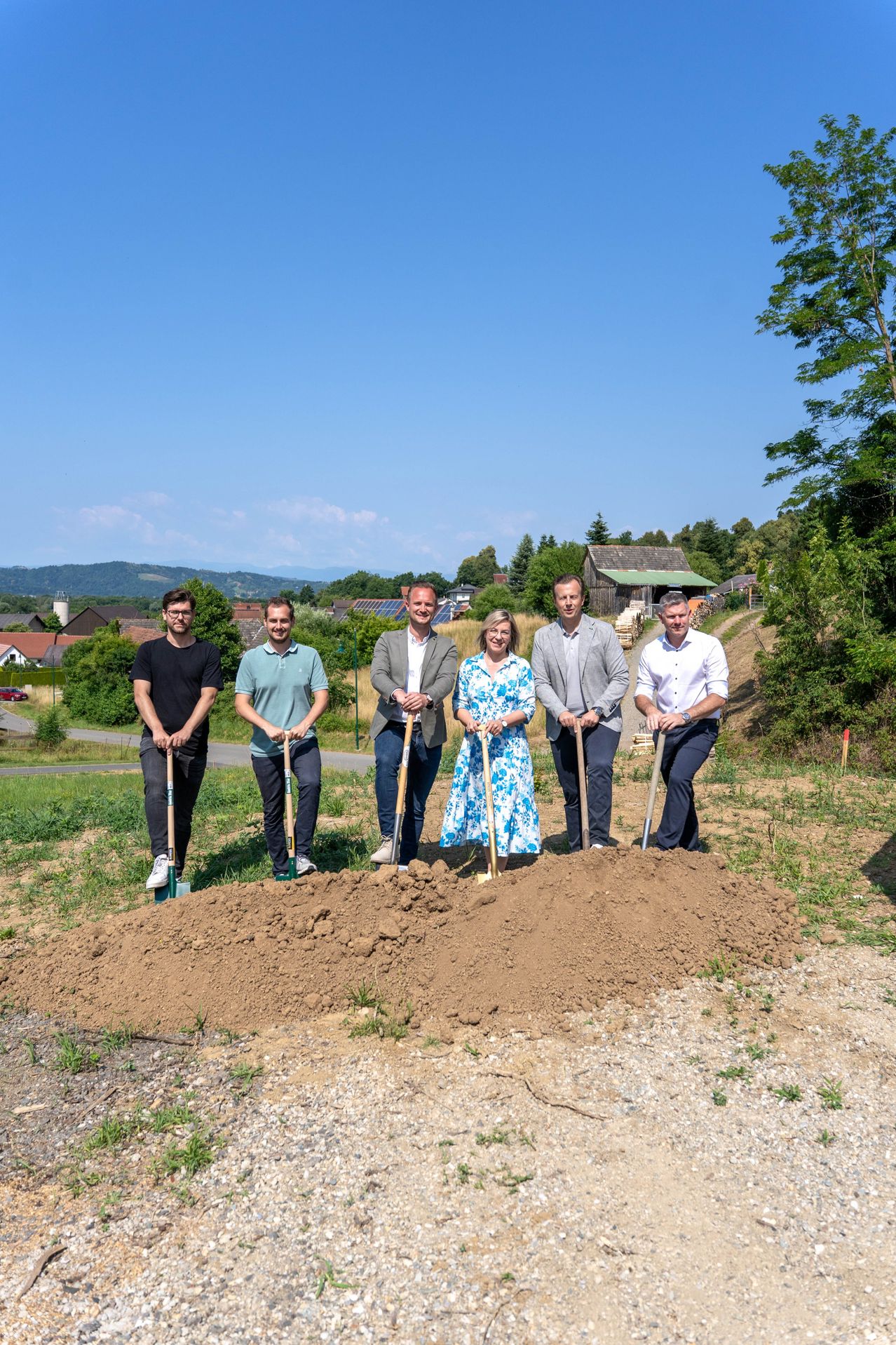 Sechs Personen stehen auf einem Erdhügel mit Schaufeln in den Händen, wahrscheinlich bei einer Grundsteinlegungsfeier. Dahinter erstreckt sich eine Landschaft mit viel Grün, entfernten Häusern und einem klaren blauen Himmel.