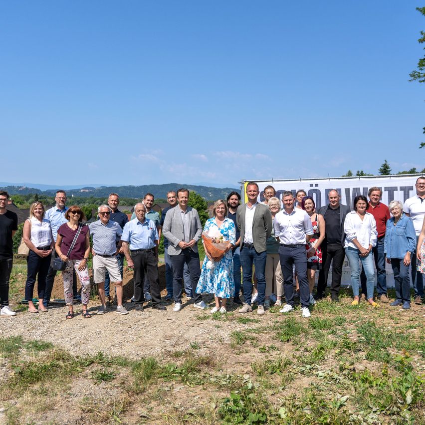 Eine Gruppe von Menschen steht vor einem großen Banner und lächelt für ein Foto. Sie befinden sich auf einem Schotterfeld mit Gras und Bergen im Hintergrund.