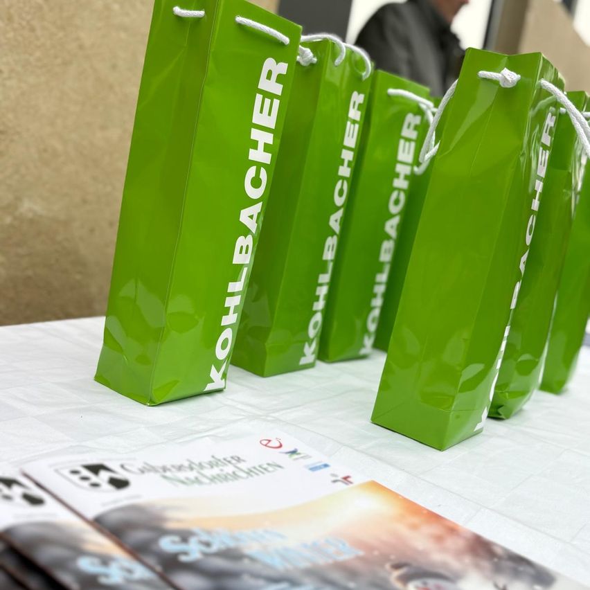 Eine Reihe grüner Geschenktüten mit der Aufschrift 'KOHLBACHER' sind auf einem Tisch ausgestellt. Auf dem Tisch liegen Broschüren und Flyer.