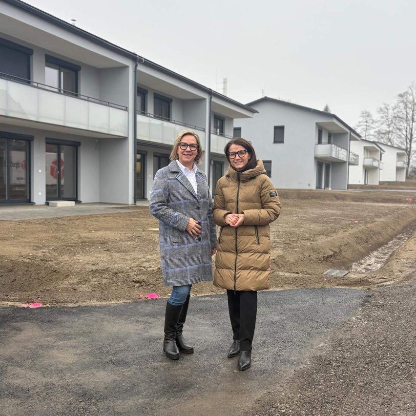 Zwei Frauen stehen vor einer Baustelle mit teilweise fertiggestellten Gebäuden. Sie lächeln und scheinen für ein Foto zu posieren.
