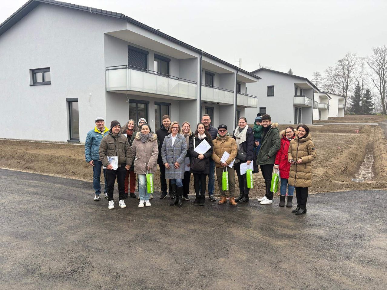 Eine Gruppe von Menschen in Winterkleidung posiert vor einem modernen Wohngebäude. Sie halten Papiere und Einkaufstüten.