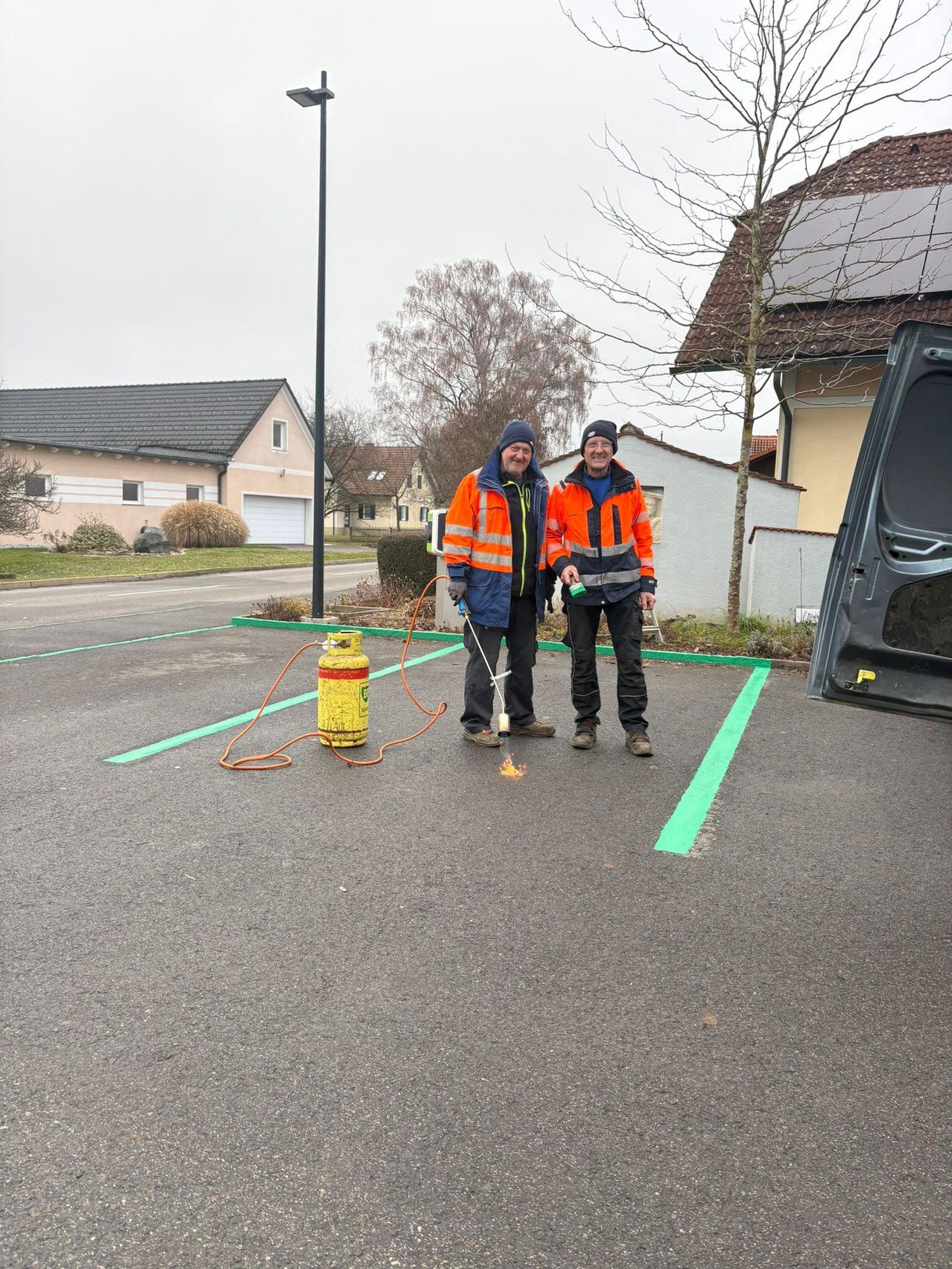 Zwei Männer in orange Sicherheitsjacken stehen auf einem Parkplatz neben einem gelben Gaszylinder. Einer von ihnen hält ein Werkzeug. Hinter ihnen befinden sich Häuser und ein Gebäude mit Solarpanelen.