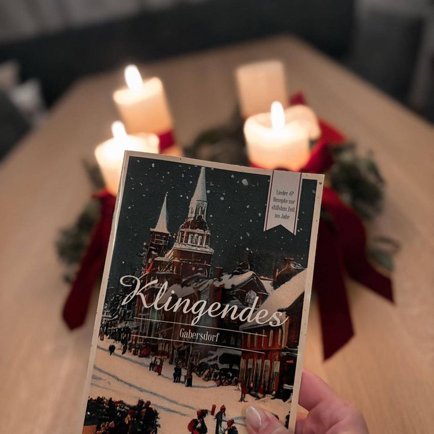 Eine Person hält ein Buch mit dem Titel Klingendes auf einem Tisch mit Kerzen und Weihnachtsdekoration.