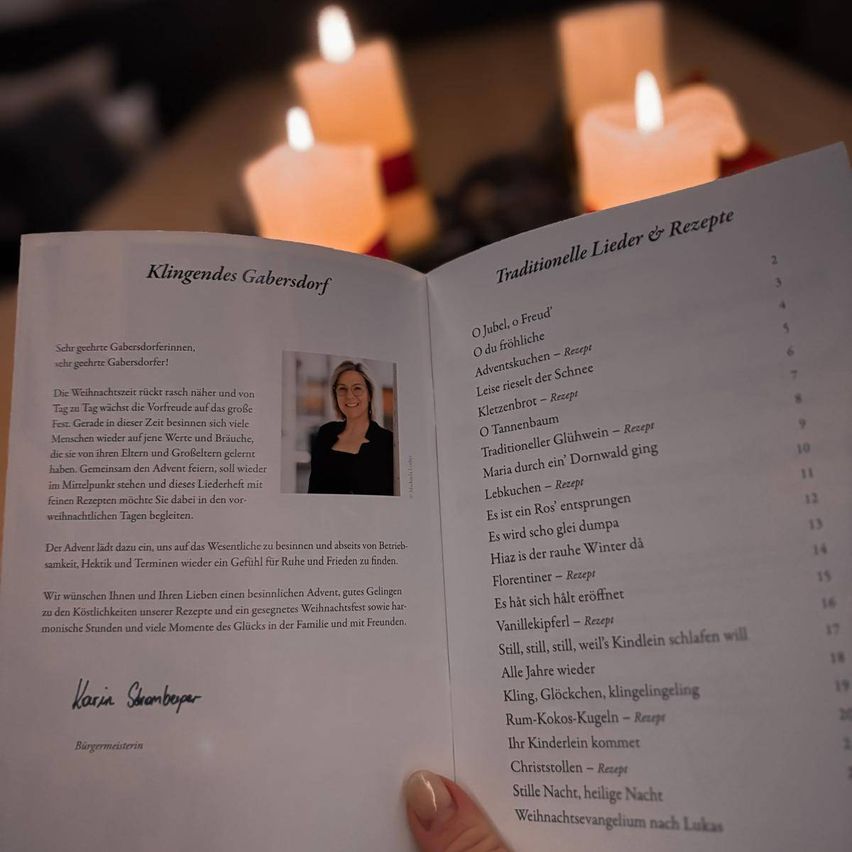 Eine Hand hält ein offenes Buch mit dem Titel 'Klingendes Gabensdorf'. Das Buch zeigt ein Foto einer Frau und Text auf Deutsch. Im Hintergrund sind verschwommene Kerzen zu sehen.
