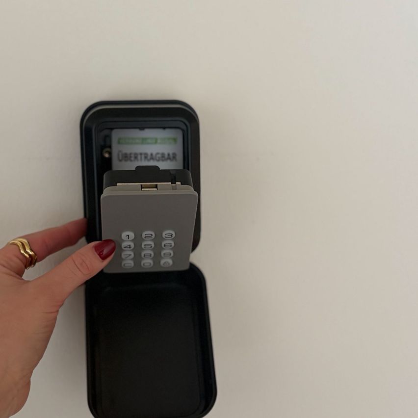 Eine Hand hält ein silbernes digitales Schloss, beschriftet mit 'Übertragbar', an einer schlichten weißen Wand. Das Schloss hat eine Tastatur mit Zahlen 1-9 und einem Schlosssymbol.