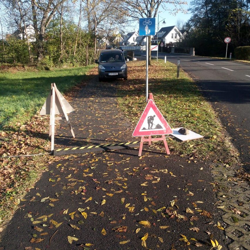 Ein Gehweg mit Bauabsperrungen und Blättern darauf, ein geparktes Auto in der Nähe und ein blaues Schild mit Fahrrad-Symbol.