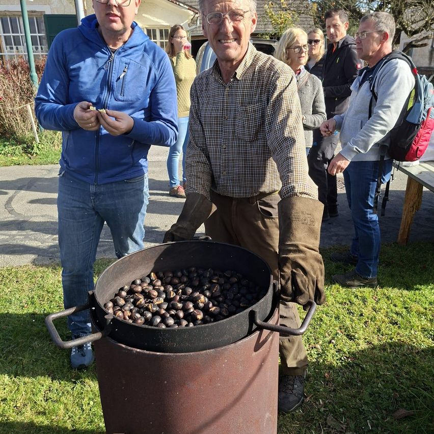 Ein älterer Mann röstet Kastanien in einem Fass, während ein Mann in einem blauen Jackett neben ihm steht. Sie befinden sich in einem Grasbereich mit mehreren Personen im Hintergrund.