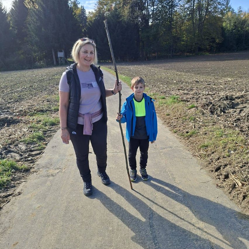 Eine Frau und ein Junge laufen lächelnd auf einem Pfad, mit einem Stock in der Hand des Jungen. Die Frau trägt eine Weste und der Junge einen Mantel. Sie sind von einem Grasfeld umgeben.