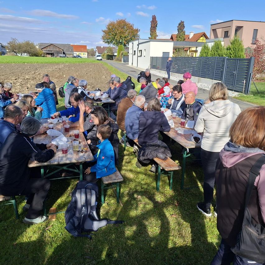 Eine Gruppe von Menschen versammelt sich um Picknicktische auf einem Grasfeld, möglicherweise für eine Gemeinschaftsveranstaltung, mit einem Zaun und Gebäuden im Hintergrund.