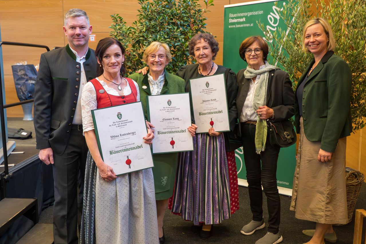 Fünf Personen posieren für ein Foto und halten Urkunden in den Händen. Eine Frau trägt ein traditionelles bayerisches Kleid. Ein grüner Banner mit dem Text 'Baeumerin' ist hinter ihnen sichtbar.