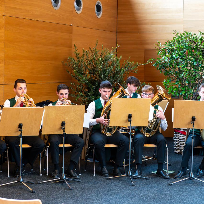 Eine Gruppe junger Musiker spielt drinnen, sitzt mit Blechblasinstrumenten vor sich. Sie spielen Trompeten und Tuben. Der Raum hat Holzwände und einen Teppichboden.