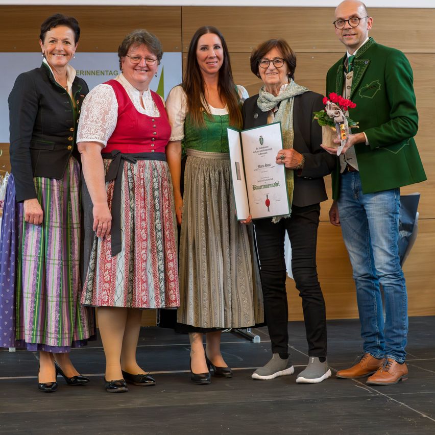 Fünf Personen stehen auf einer Bühne in traditioneller Kleidung. Eine Frau hält ein Zertifikat und Blumen. Ein Mann in Grün schaut sie lächelnd an. Hinter ihnen steht ein Banner mit der Aufschrift 'Erinnerntag'.