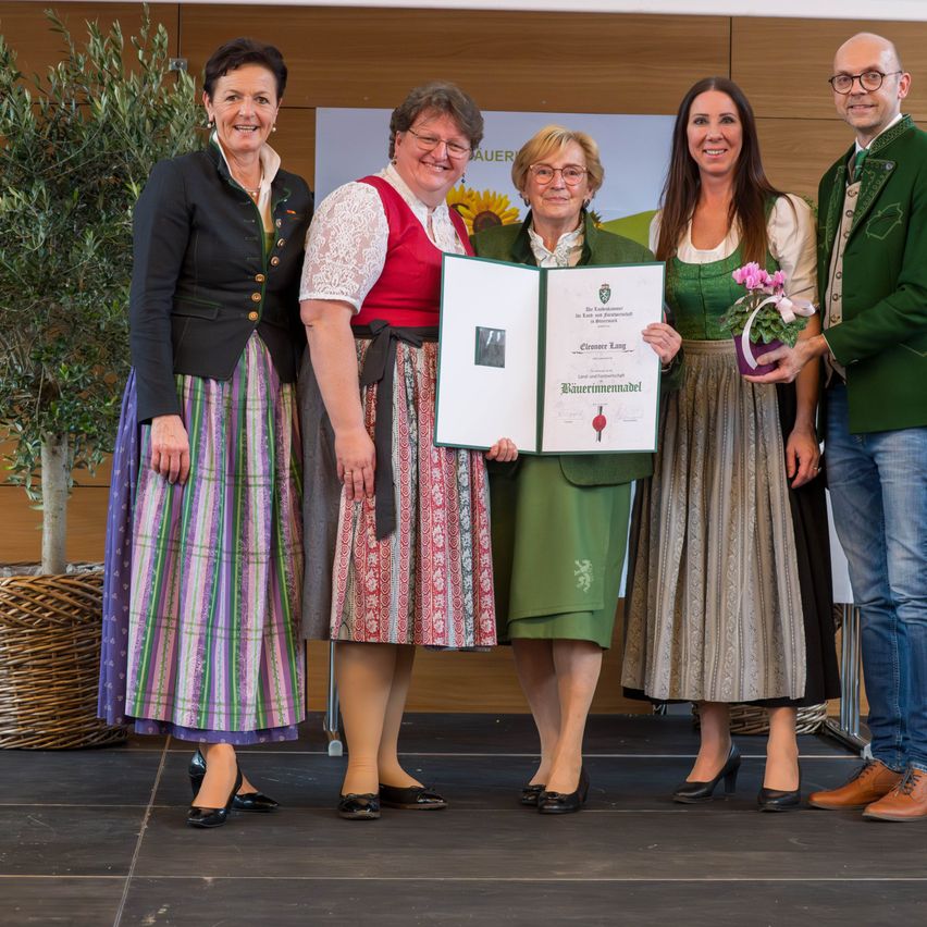 Fünf Personen stehen auf einer Bühne, eine Frau hält ein Zertifikat und einen Blumenstrauß. Die Frau in der Mitte hält das Zertifikat, und die Frau auf der rechten Seite hält einen Blumenstrauß. Die anderen drei stehen dahinter. Dahinter steht eine Topfpflanze.