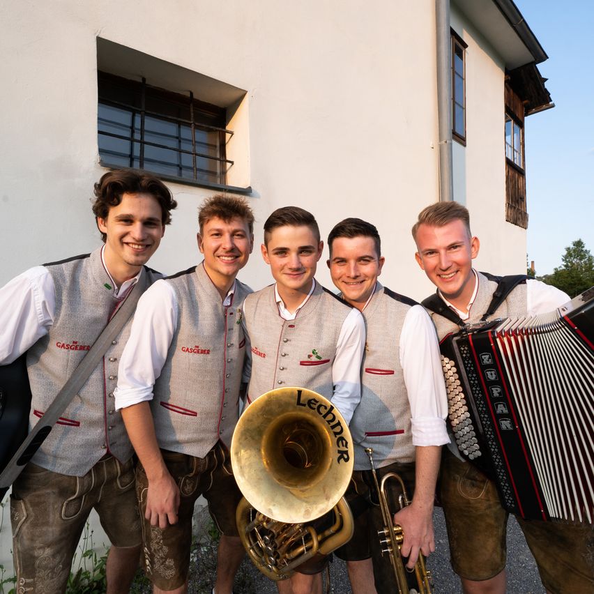 Fünf junge Männer in traditioneller deutscher Kleidung, lächelnd, posieren für ein Foto vor einem Gebäude. Sie halten Musikinstrumente, darunter Trompete, Posaune und Akkordeon.