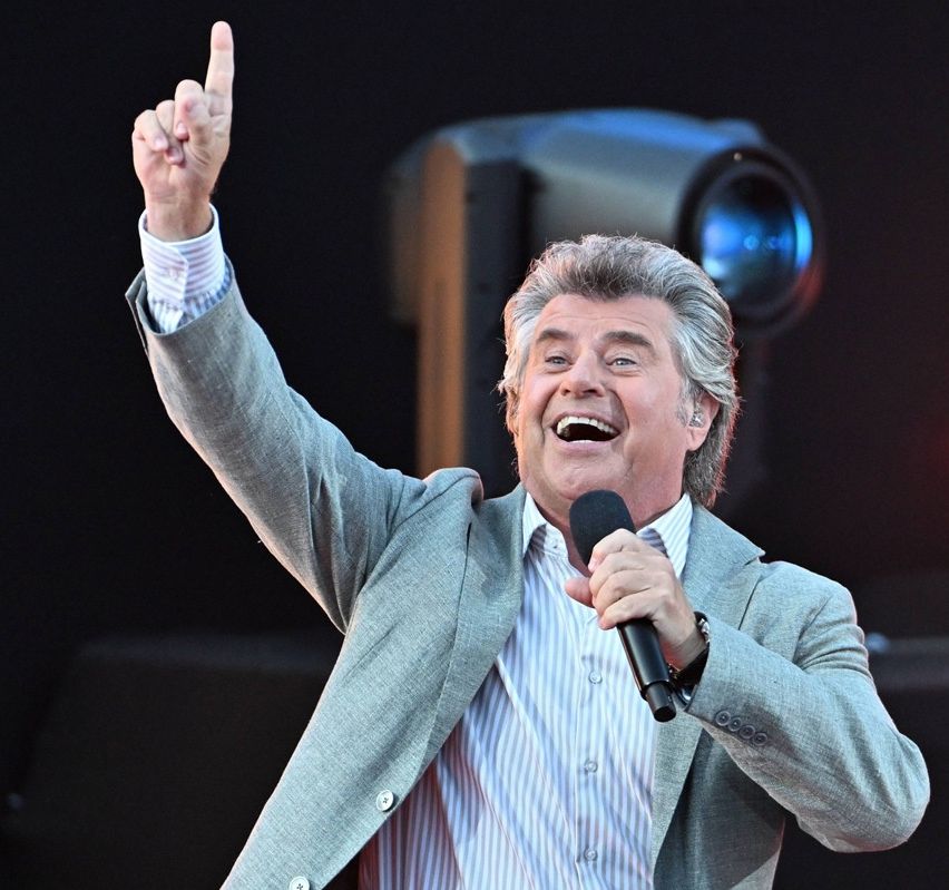 Ein Mann in einem grauen Anzug und gestreiftem Hemd lächelt und hält ein Mikrofon auf der Bühne.