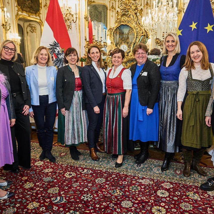 A group of women in traditional attire stand in a formal setting, smiling for a photograph with flags behind them.