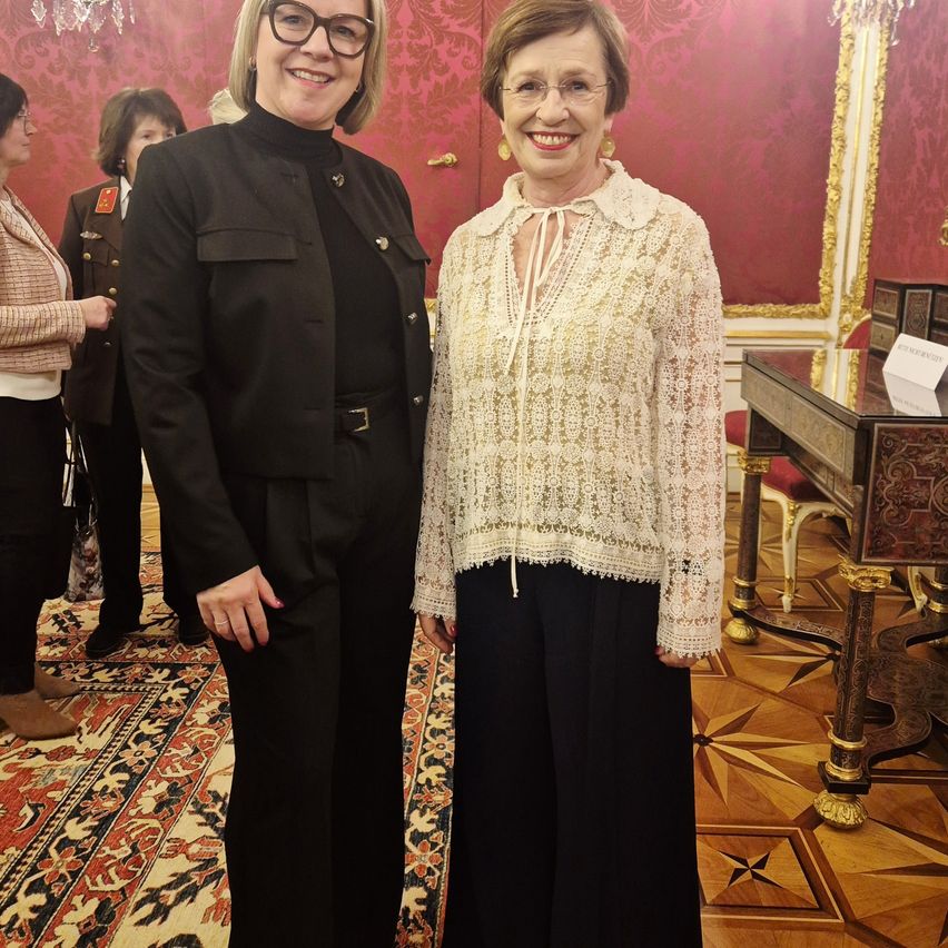 Two women stand in a room with ornate red walls. One woman in glasses and a black coat smiles. The other wears a lace blouse and black pants. Behind them, a desk is adorned with intricate designs.